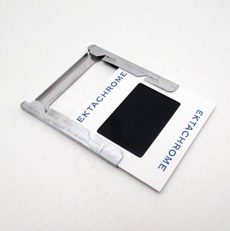 Pu&ograve; includere: Un supporto per diapositive bianco con la scritta "Ektachrome" in blu, che contiene una diapositiva nera. Il supporto &egrave; tenuto in posizione da un supporto per diapositive in metallo argentato.