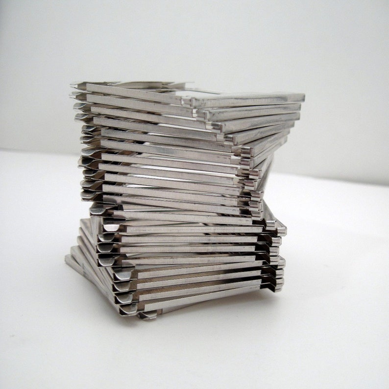 Pu&ograve; includere: Una pila di forcine per capelli in metallo argentato disposte a spirale. Le forcine sono rettangolari con una leggera curva e sono impilate per creare un'unica scultura astratta. Lo sfondo &egrave; bianco.