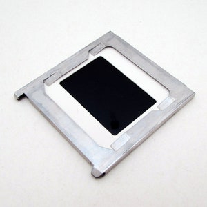 Pu&ograve; includere: Un supporto per diapositive quadrato, color argento, con un'apertura rettangolare nera al centro. Il supporto ha un bordo bianco attorno all'area nera ed &egrave; progettato per contenere una diapositiva fotografica. Il supporto &egrave; su una superficie bianca.