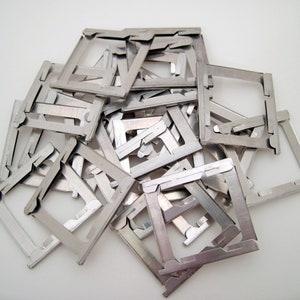 Pu&ograve; includere: Un mucchio di piccoli telai metallici color argento, ciascuno con una forma quadrata e un design dentellato unico. I telai sono sparsi su una superficie bianca, creando un'estetica industriale strutturata.
