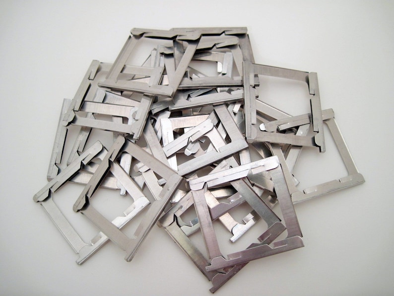 Pu&ograve; includere: Un mucchio di piccoli telai metallici color argento, ciascuno con una forma quadrata e un design dentellato unico. I telai sono sparsi su una superficie bianca, creando un'estetica industriale strutturata.
