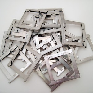 以下が含まれることがあります： 正方形の形状とユニークなデザインを持つ、小さな銀色の金属フレームの山。フレームは白い表面に散らばっており、質感のある金属的な表示になっています。フレームは、より大きなアイテムのコンポーネントのようです。