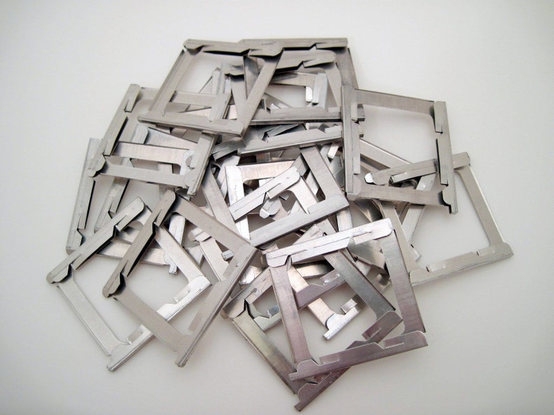 May include: A pile of small, silver-colored metal frames, each with a square shape and a unique design. The frames are scattered on a white surface, creating a textured, metallic display. The frames appear to be components of a larger item.