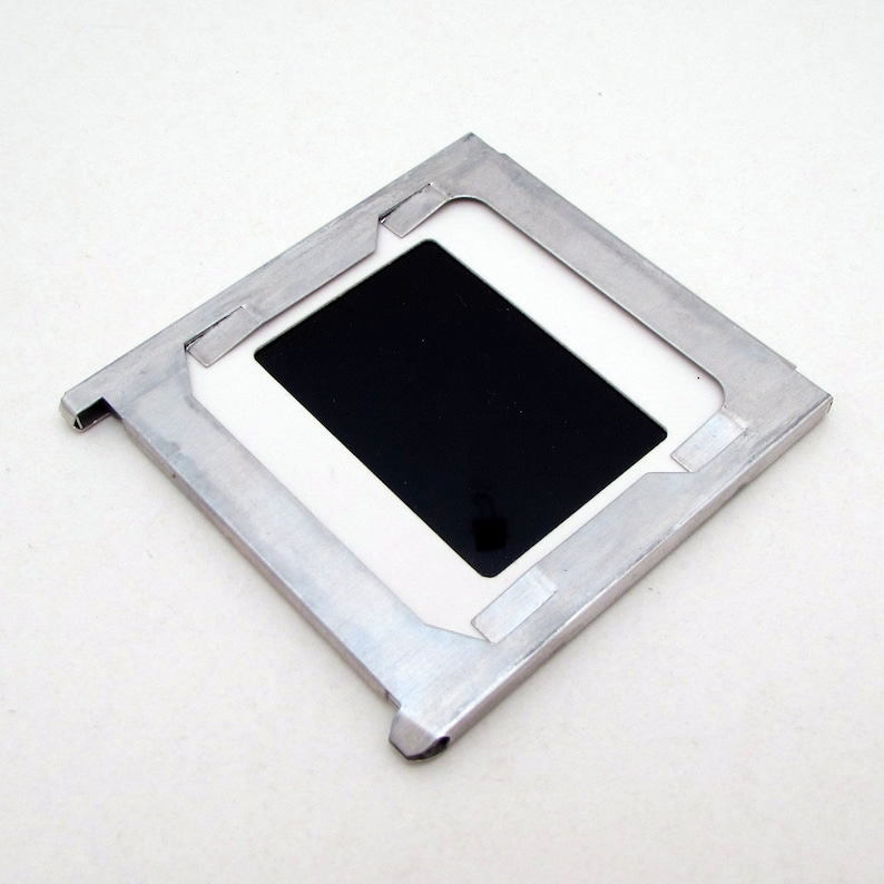 May include: A square, silver-colored slide mount with a black rectangular insert. The mount has a white border around the black insert and is made of a sturdy metal. The slide mount is designed to hold a photographic slide.