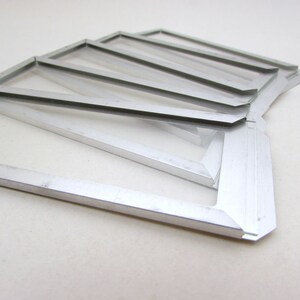 May include: A collection of silver-colored, rectangular frames with clear glass inserts. The frames are arranged in a fan-like pattern, with a few overlapping. The edges of the frames are clean and sharp, and the overall aesthetic is modern and minimalist.