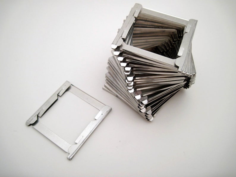 Pu&ograve; includere: Una collezione di supporti per diapositive in metallo color argento, uno piatto e gli altri impilati a spirale. I supporti sono rettangolari con una finestra trasparente, progettati per diapositive fotografiche. Lo sfondo &egrave; bianco.