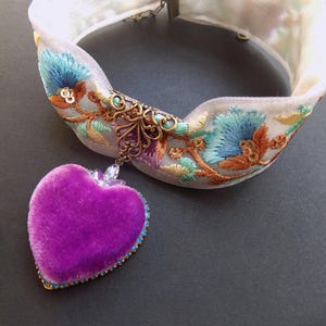 May include: A white fabric choker necklace with embroidered blue flowers and orange leaves. A purple velvet heart pendant with a blue gemstone border hangs from the center. The necklace has a decorative metal clasp.