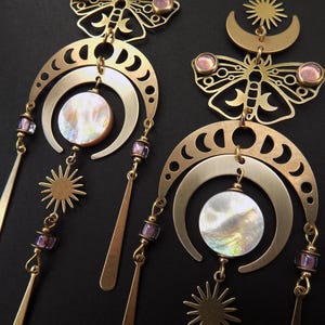 May include: A pair of gold-toned metal earrings featuring a moth silhouette at the top, a crescent moon phase arch, a central iridescent circular shell inlay, a sunburst charm, and dangling metal bars accented with small purple glass beads.