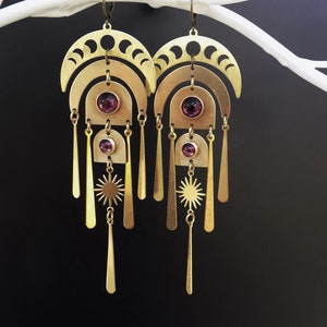 May include: A pair of brass-colored earrings with a celestial design. Each earring features crescent moon shapes, arching forms, a sunburst, and dangling bars. Round, purple gemstones are set in the center of each earring. The earrings hang from a simple hook.