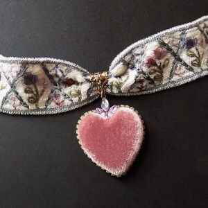 A white embroidered choker necklace with a pink velvet heart pendant. The choker features floral embroidery in shades of pink, purple, and green. The heart pendant is trimmed with crystals