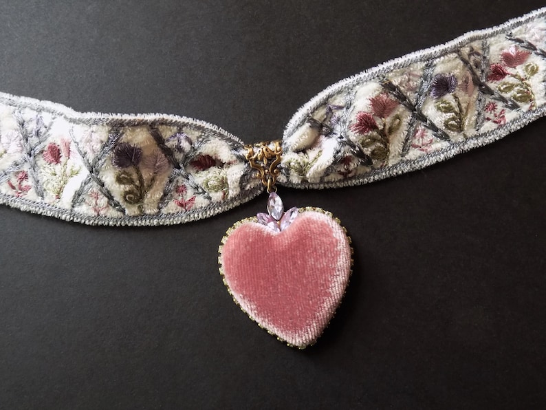 A white embroidered choker necklace with a pink velvet heart pendant. The choker features floral embroidery in shades of pink, purple, and green. The heart pendant is trimmed with crystals