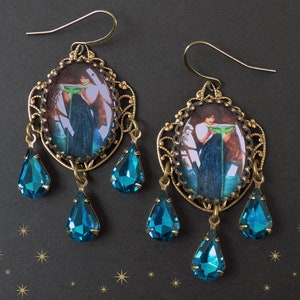 May include: A pair of gold-tone earrings with a blue glass teardrop design. Each earring features a round, oval-shaped pendant with a vintage-style image of a woman holding a green leaf. The pendants are surrounded by a delicate filigree design.