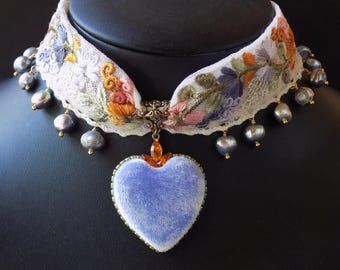 Puede incluir: Un collar tipo gargantilla con una banda de encaje blanco bordada con motivos florales coloridos en tonos naranja, rosa y morado. La banda está adornada con perlas grises colgantes y un colgante central en forma de corazón de terciopelo azul claro.