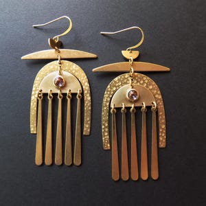 May include: A pair of gold-toned dangle earrings featuring a horizontal bar, a textured arch, and a semi-circular piece with a small purple stone. Five thin, flat metal strips hang from the base of each earring, suspended by gold-toned ear hooks.