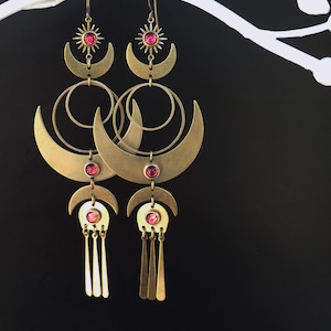 May include: A pair of long, brass-colored earrings with a celestial theme. Each earring features a sun, crescent moons, and circular elements, adorned with red gemstones. The earrings have a dangling design with vertical bars.