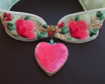 Puede incluir: Un collar verde menta con flores rojas bordadas y hojas verdes. Un colgante en forma de corazón rosa con ribete dorado y detalles azules cuelga del centro. El collar está hecho de una tela transparente.