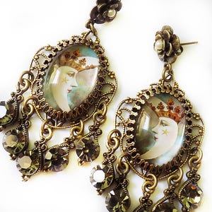 May include: Antique-style earrings with a celestial theme. Each earring features an oval pendant with a moon face design, framed by ornate filigree and dangling teardrop-shaped crystals. The earrings have a bronze finish.