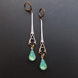 May include: A pair of long drop earrings featuring bronze-toned leverback closures and slender, vertical metal bars. Each earring includes an openwork geometric frame that connects to a faceted, pear-shaped mint green crystal pendant at the base.