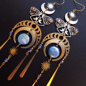 May include: A pair of long gold-toned earrings featuring a moth silhouette at the top, a crescent moon phase arch, and a central iridescent blue circular cabochon. The design includes dangling sunburst charms, small purple beads, and thin metal spikes.