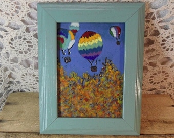May include: A framed artwork featuring a blue sky with colorful hot air balloons. The balloons are in various colors, including rainbow stripes. The bottom of the artwork depicts a colorful, textured landscape. The frame is a light blue color.