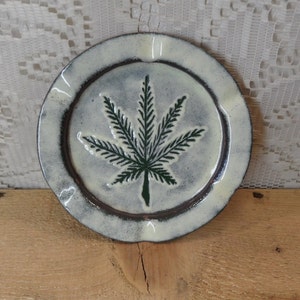 May include: A white ceramic ashtray with a green marijuana leaf design. The ashtray has a slightly raised rim and a distressed finish.