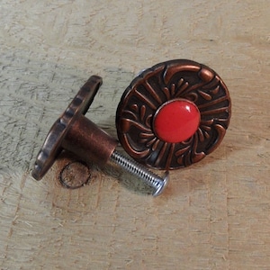 May include: Two decorative cabinet knobs with a copper finish and a red center. The knobs have an ornate, floral design around the red center. One knob is lying on its side, showing the screw.