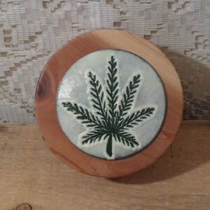 May include: A circular wooden coaster featuring a central inset ceramic disc. The disc is off-white with a dark green embossed cannabis leaf design. The wood has a natural light brown grain with visible knots, creating a contrast with the smooth, glazed finish of the central ceramic inlay.