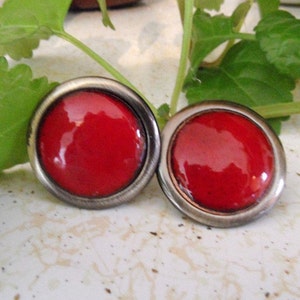 May include: A pair of round, red earrings with a silver-colored metal border. The earrings have a glossy finish and are set against a background of green leaves and a light-colored surface. The earrings are a classic accessory.
