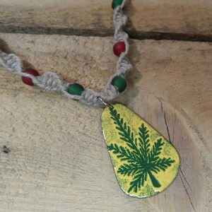 May include: A hemp necklace with green and red beads and a pendant featuring a green fern leaf on a yellow background.