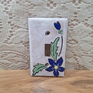 May include: A ceramic light switch cover with a floral design. The cover is rectangular with a white background and a blue flower and green leaves. The switch opening is rectangular.