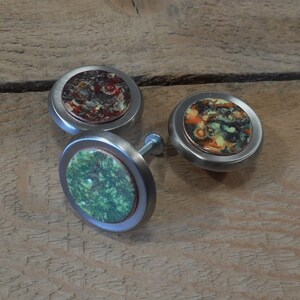 May include: Three round cabinet knobs with a silver metal base and a colorful ceramic top. The knobs feature abstract patterns in shades of green, orange, and brown.