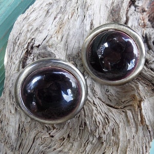 May include: A pair of round, dark purple glass earrings with silver-toned metal settings. The earrings have a smooth, glossy finish.