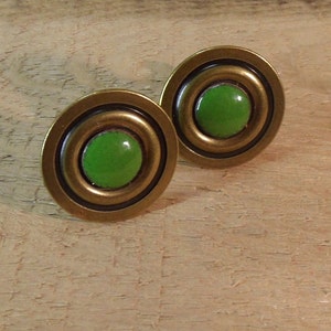 May include: A pair of round cufflinks with a brass-colored metal frame. Each cufflink features a bright green, circular center. The design includes concentric circles, creating a layered effect. The cufflinks are displayed on a light brown wooden surface.