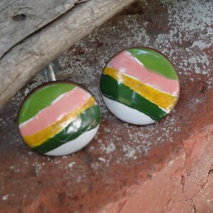 May include: Two round, colorful cabinet knobs. Each knob features a design with green, pink, and yellow stripes over a white base. The knobs are set against a textured, reddish-brown brick surface.