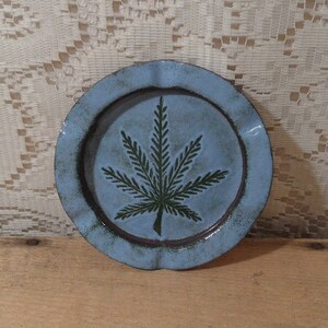 May include: A round, light blue ceramic plate with a dark green cannabis leaf design in the center. The plate has a wavy edge and a dark brown rim. The plate is sitting on a wooden surface.