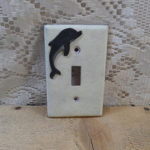 May include: A white ceramic light switch cover with a black dolphin silhouette. The dolphin is jumping out of the water.