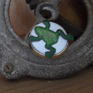 May include: A round, gold-rimmed button with a white background and a green frog design. The frog has a textured, speckled appearance. The button is set against a blurred, textured background.