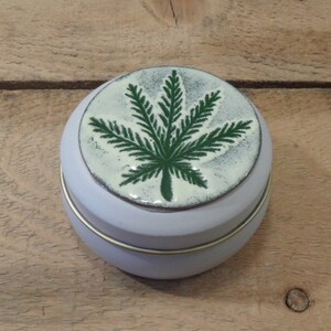 May include: A small, round, light purple tin with a lid featuring a green cannabis leaf design. The leaf is centered on a white background with a speckled texture. The tin has a gold-colored band around the middle.