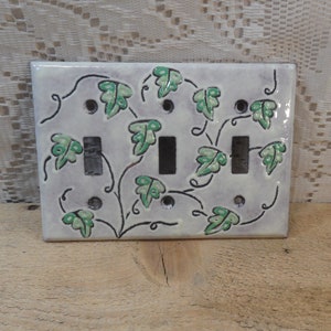 May include: A white ceramic light switch cover with a green ivy vine design. The switch cover has two toggle switches.