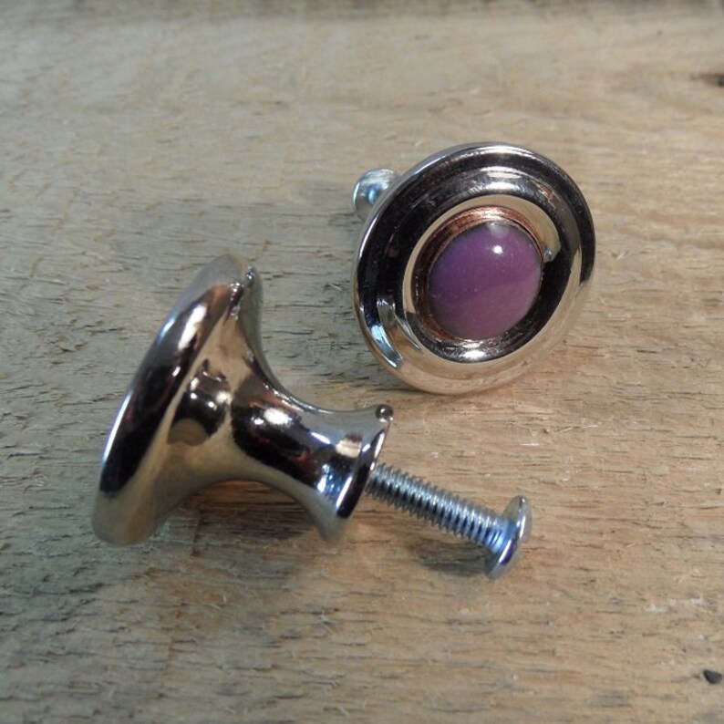 Peut inclure: Deux boutons de meuble argent&eacute;s avec une pierre centrale violette. Les boutons ont un design arrondi avec une vis pour l'installation. Les boutons sont sur une surface en bois brun clair.