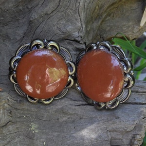 May include: A pair of vintage clip-on earrings. Each earring features a glossy, reddish-brown, oval-shaped center stone, surrounded by a decorative, silver-toned metal frame. The frames have a scalloped edge design.