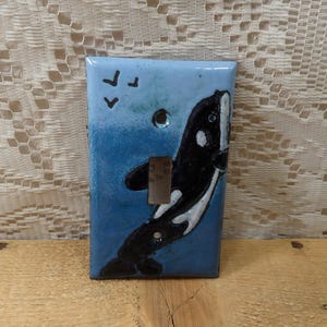 May include: A ceramic light switch cover with a blue and white background featuring a black and white orca whale jumping out of the water. Three small black birds are flying above the orca.