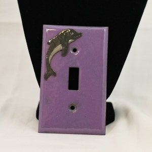 May include: A lavender light switch cover with a silver dolphin accent. The dolphin is positioned in the upper left corner. The switch plate has a rectangular opening for the switch and two holes for screws.