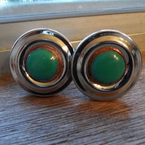 May include: A pair of round cufflinks with a shiny silver-colored outer ring, a copper-colored middle ring, and a bright green center. The cufflinks are displayed on a wooden surface.