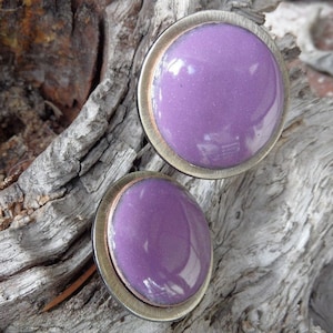 May include: A pair of round stud earrings with a shiny, light purple center. The earrings have a silver-colored metal frame with a copper-colored inner ring. The earrings are displayed on a piece of weathered wood.