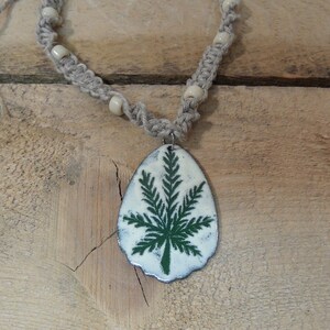 May include: A handmade necklace featuring a pendant with a green leaf design on a white background. The pendant is suspended from a braided hemp cord with small, off-white beads. The necklace rests on a wooden surface.