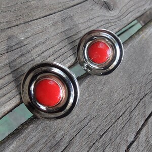 May include: A pair of vintage silver-toned earrings with a concentric design. Each earring features a bright red, circular center surrounded by multiple silver rings. The earrings are set against a weathered wooden surface.