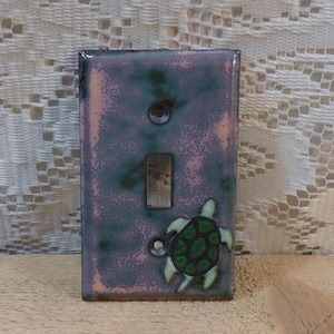 May include: A light switch cover with a pink and green glaze. A small sea turtle is painted on the cover.
