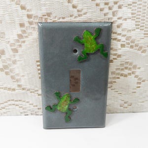 May include: A gray ceramic light switch cover with two green frog designs. The frogs have a textured appearance and are positioned as if climbing. The switch plate has a rectangular wooden switch. The background is a white lace fabric.