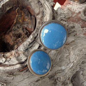 May include: A pair of round, blue enamel earrings with a silver-colored border. The earrings are set against a textured, weathered wood background, creating a contrast of textures and colors. The earrings are a simple, yet elegant accessory.